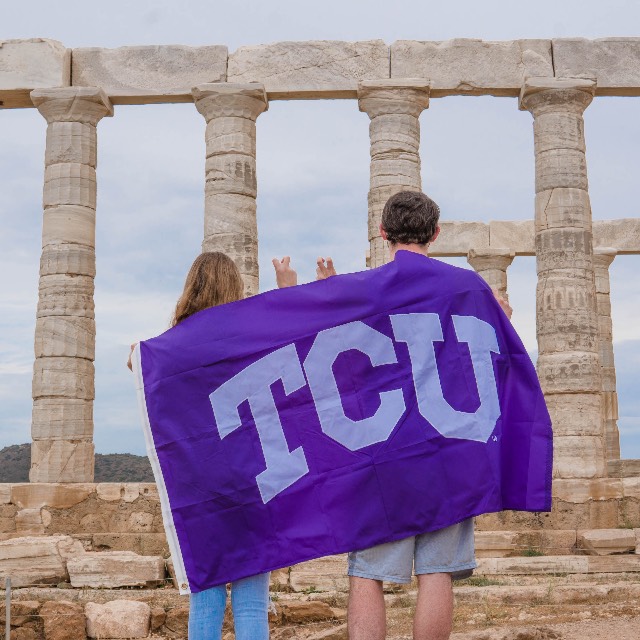 Two students, backs toward us, drape a TCU flag across their shoulders as they look at the pillars of a classical monument