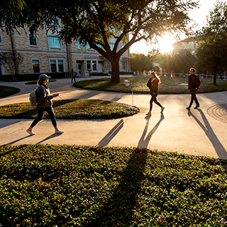 TCU students wearing jackets walk on campus, casting long shadows on a sunny day