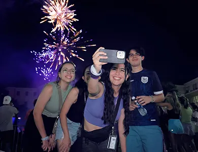 Four students gather for a selfie as colorful fireworks explode in the background