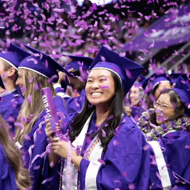Graduates in purple caps and gowns celebrate during a TCU commencement ceremony as purple confetti falls, with a smiling graduate holding a confetti popper in the foreground.