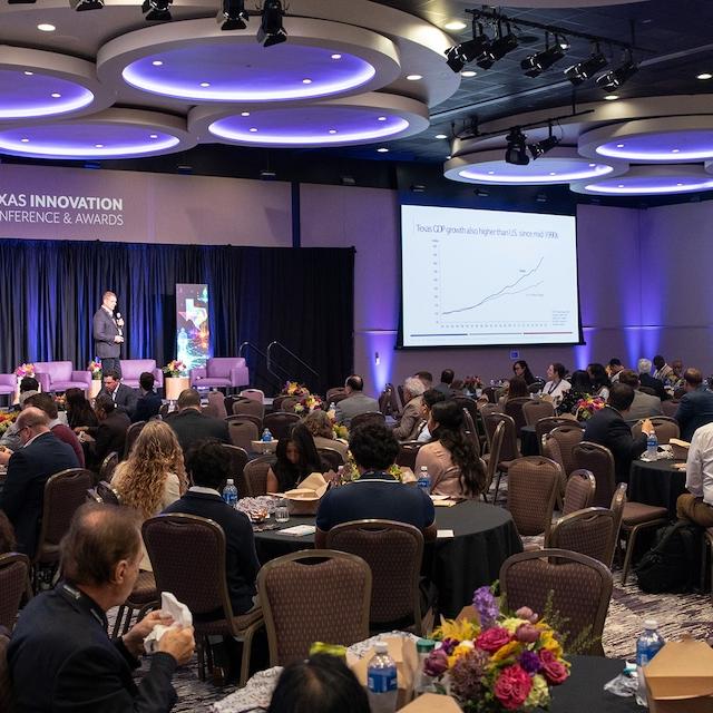 Image of people sitting at tables in a large banquet hall with a speaker on stage